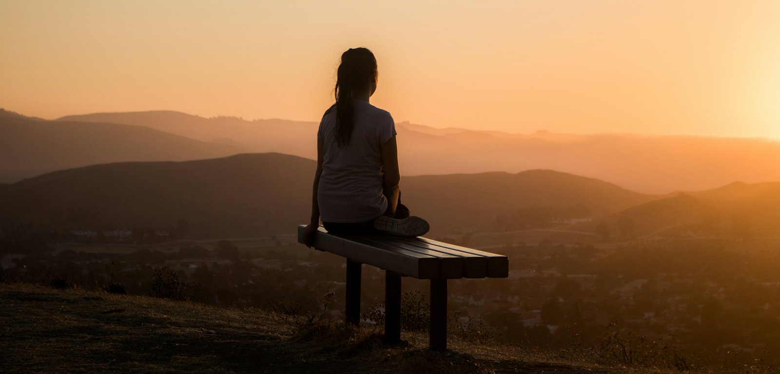 A woman sitting on a bench looking out over mountains at sunset